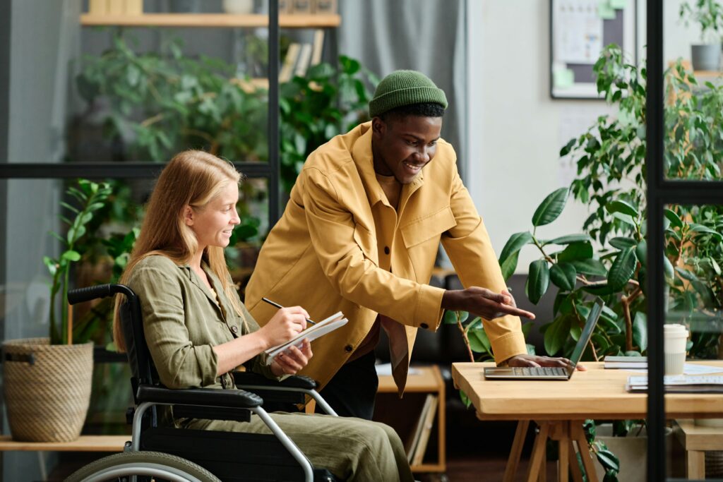 A man showing a young woman in a wheelchair accessibility software tools on a laptop device while she takes notes.